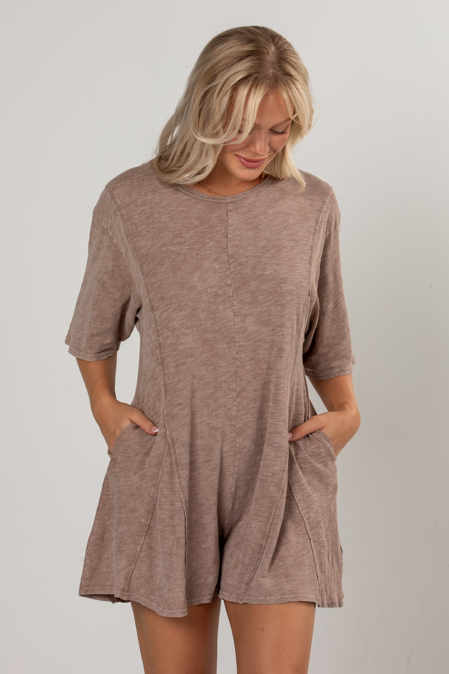 A woman wearing a short-sleeve, loose-fitting romper in a heathered taupe-brown color, standing against a light gray studio background.
