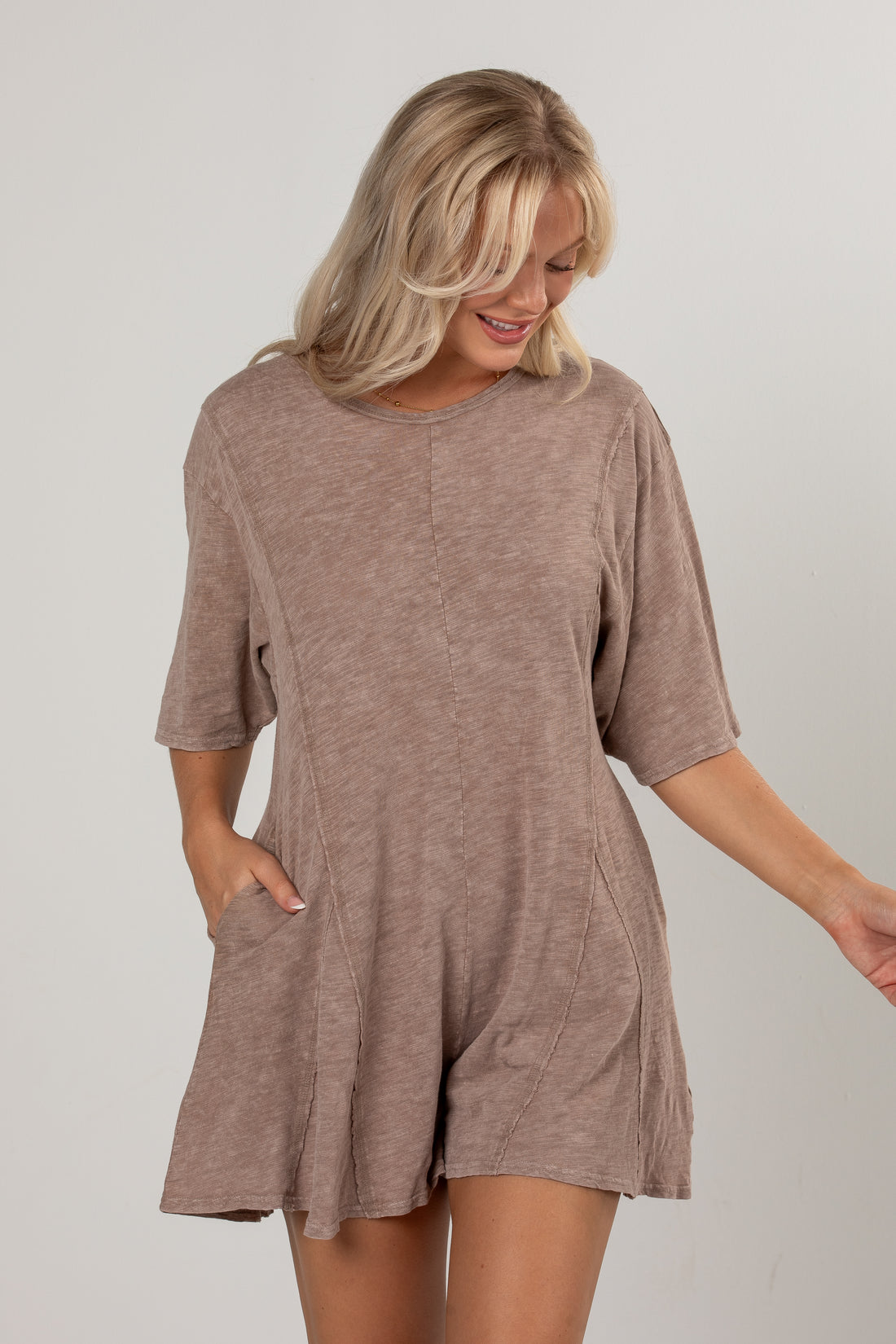 A woman wearing a short-sleeve, loose-fitting romper in a heathered taupe-brown color, standing against a plain light gray studio background.