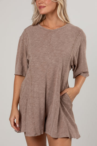 A woman wearing a loose, short-sleeve taupe-brown romper with side pockets, shown from the neck down against a light gray studio background.
