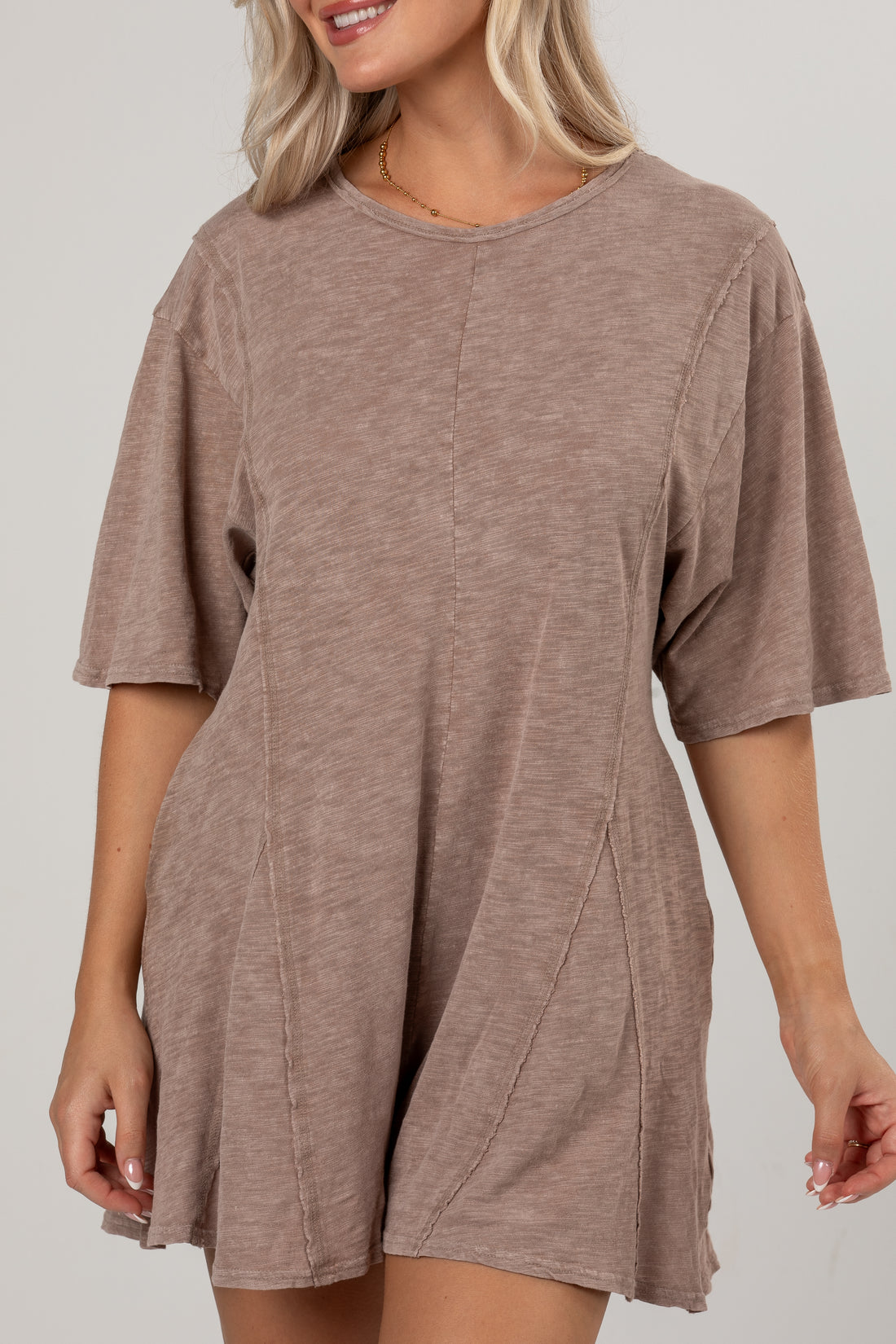 A woman wearing a loose, short-sleeve romper in a heathered taupe-brown color, shown from the waist up against a plain light gray studio background.