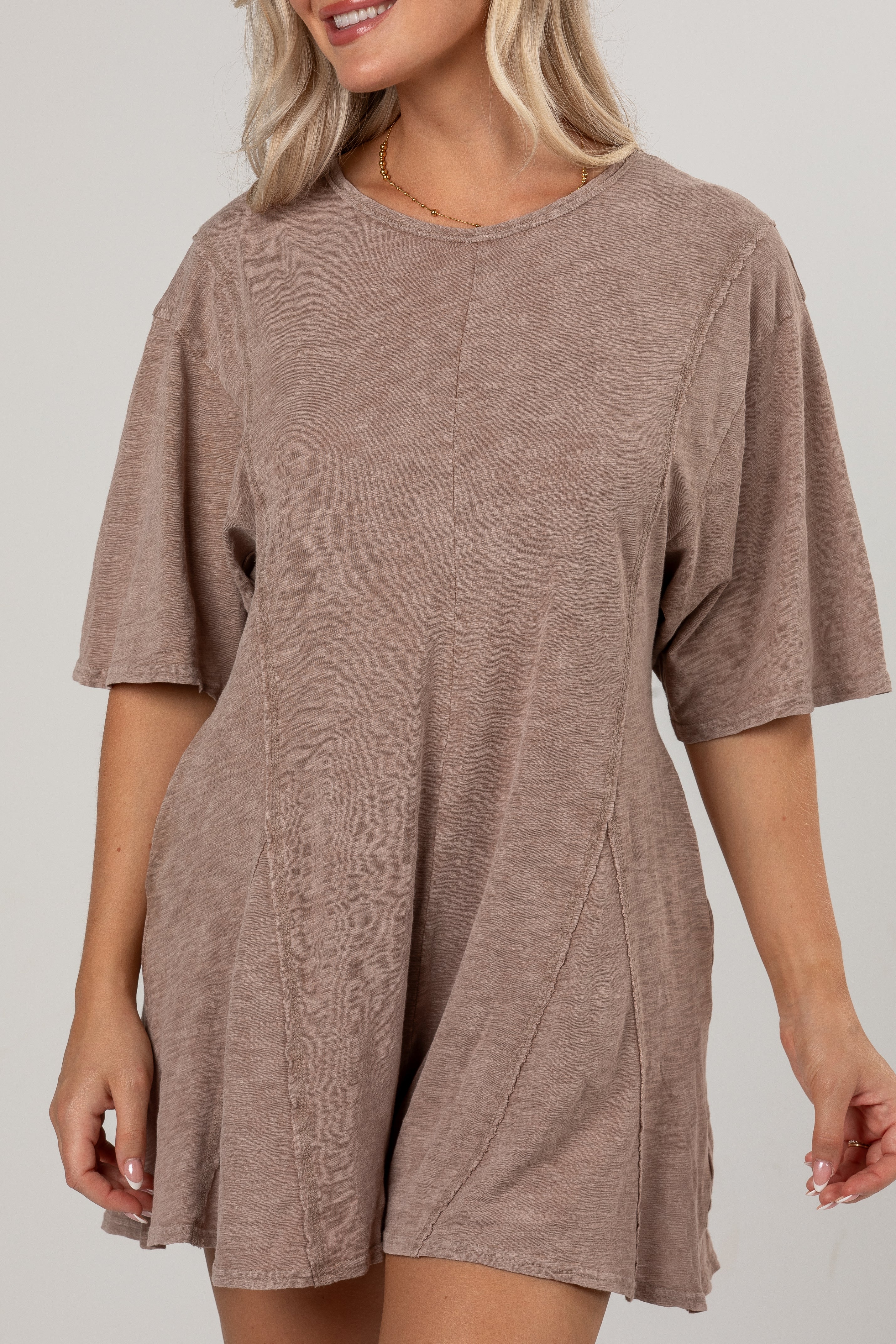 A woman wearing a loose, short-sleeve romper in a heathered taupe-brown color, shown from the waist up against a plain light gray studio background.