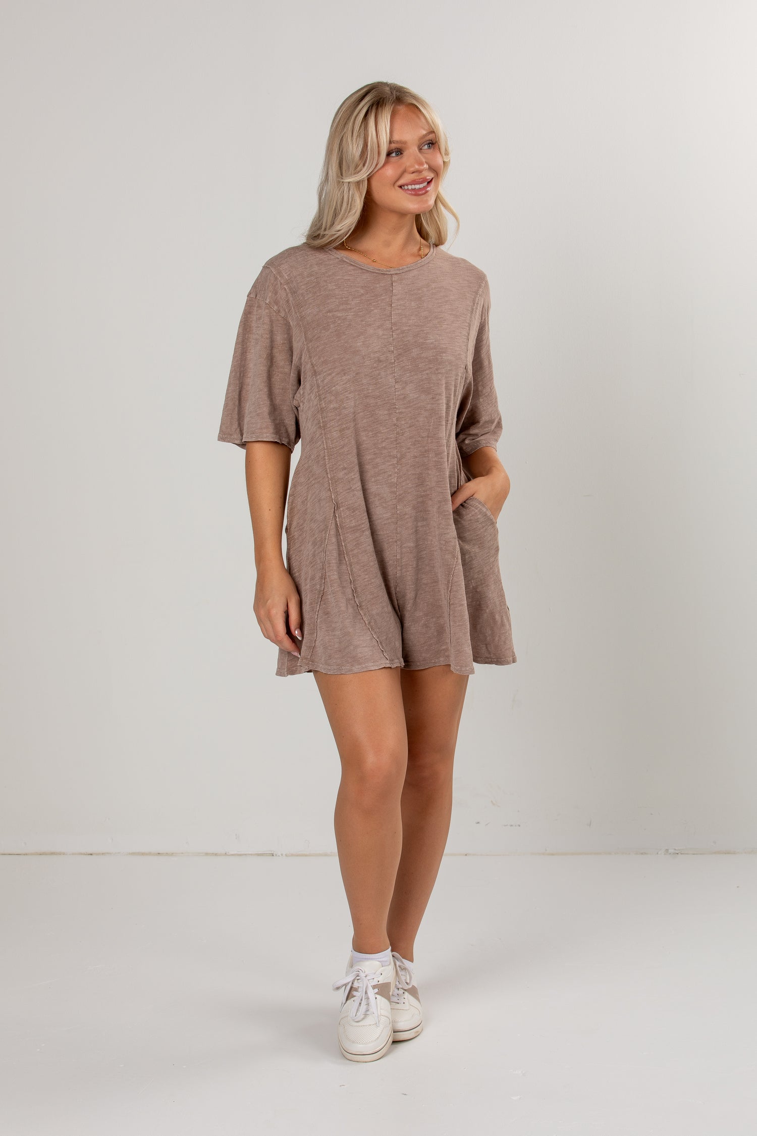 A woman modeling a short-sleeve, loose-fit mini romper in a heathered taupe-brown color, paired with white sneakers, against a plain white studio background.