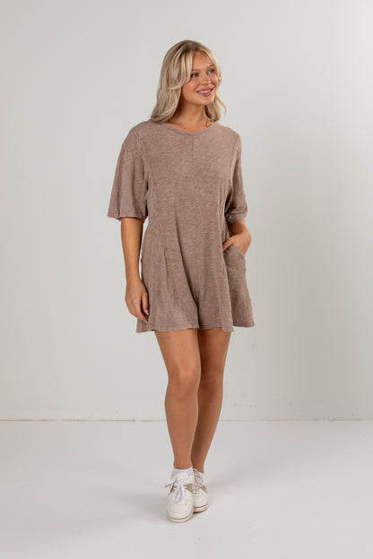 A woman modeling a short-sleeve, loose-fit mini romper in a heathered taupe-brown color, paired with white sneakers, against a plain white studio background.