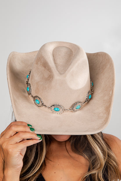 Beige cowboy hat with turquoise and silver decorative band held by a person against a neutral background
