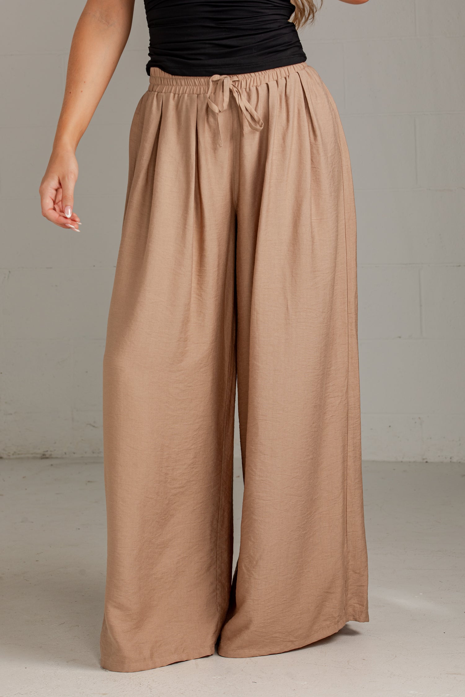 A person wearing loose, wide-leg tan pants with an elastic drawstring waist, paired with a black sleeveless top, standing against a light gray wall.