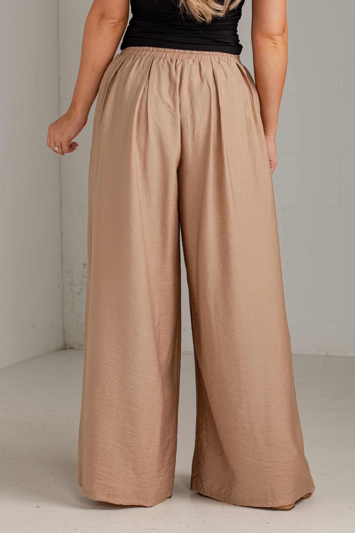 A person wearing loose, wide-leg pleated pants in a warm taupe-beige color, paired with a black sleeveless top. The photo is taken from the back in a simple studio setting with a light gray wall and floor.