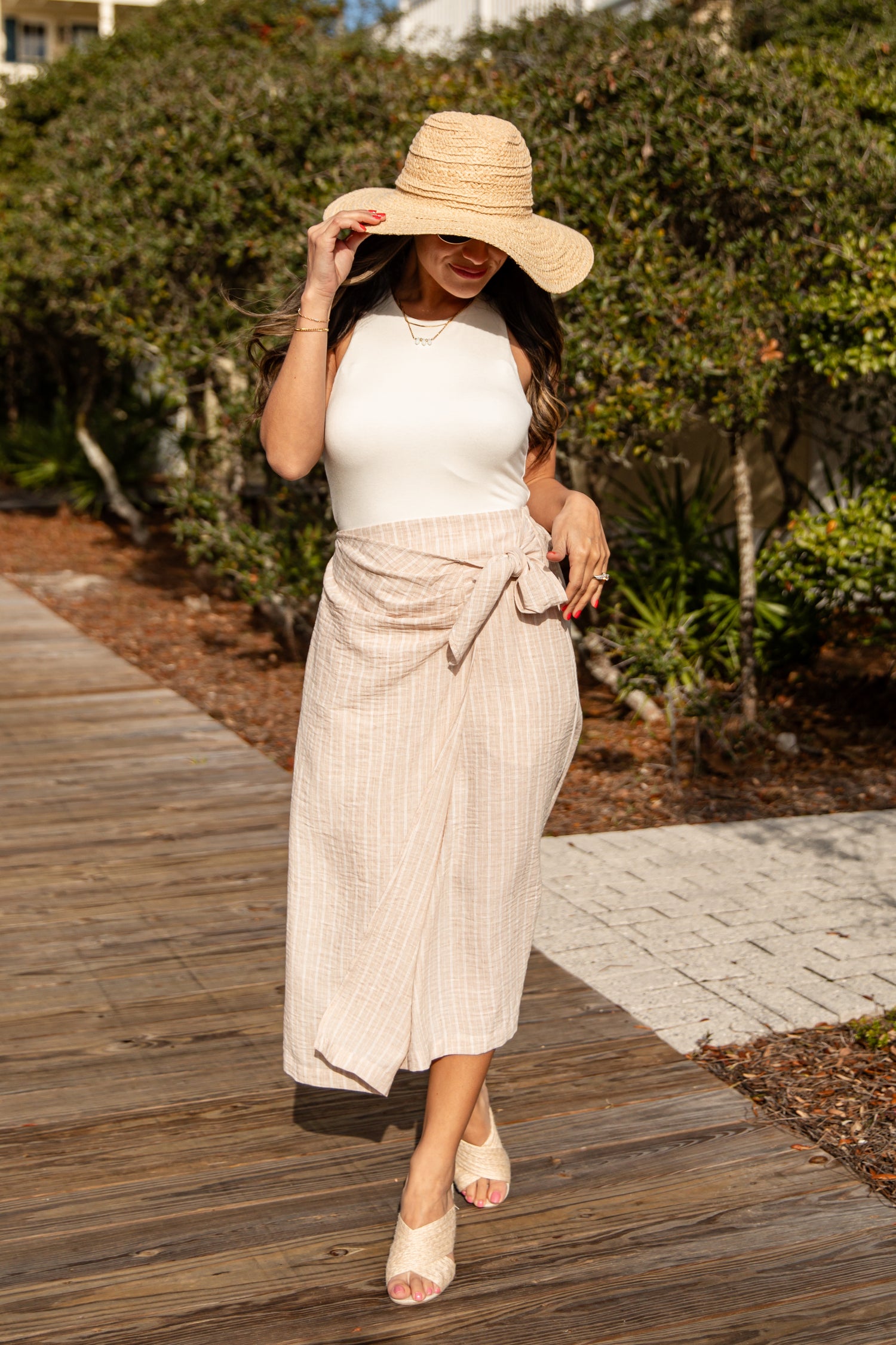 Woman wearing a white top and beige skirt with a straw hat on a wooden path.