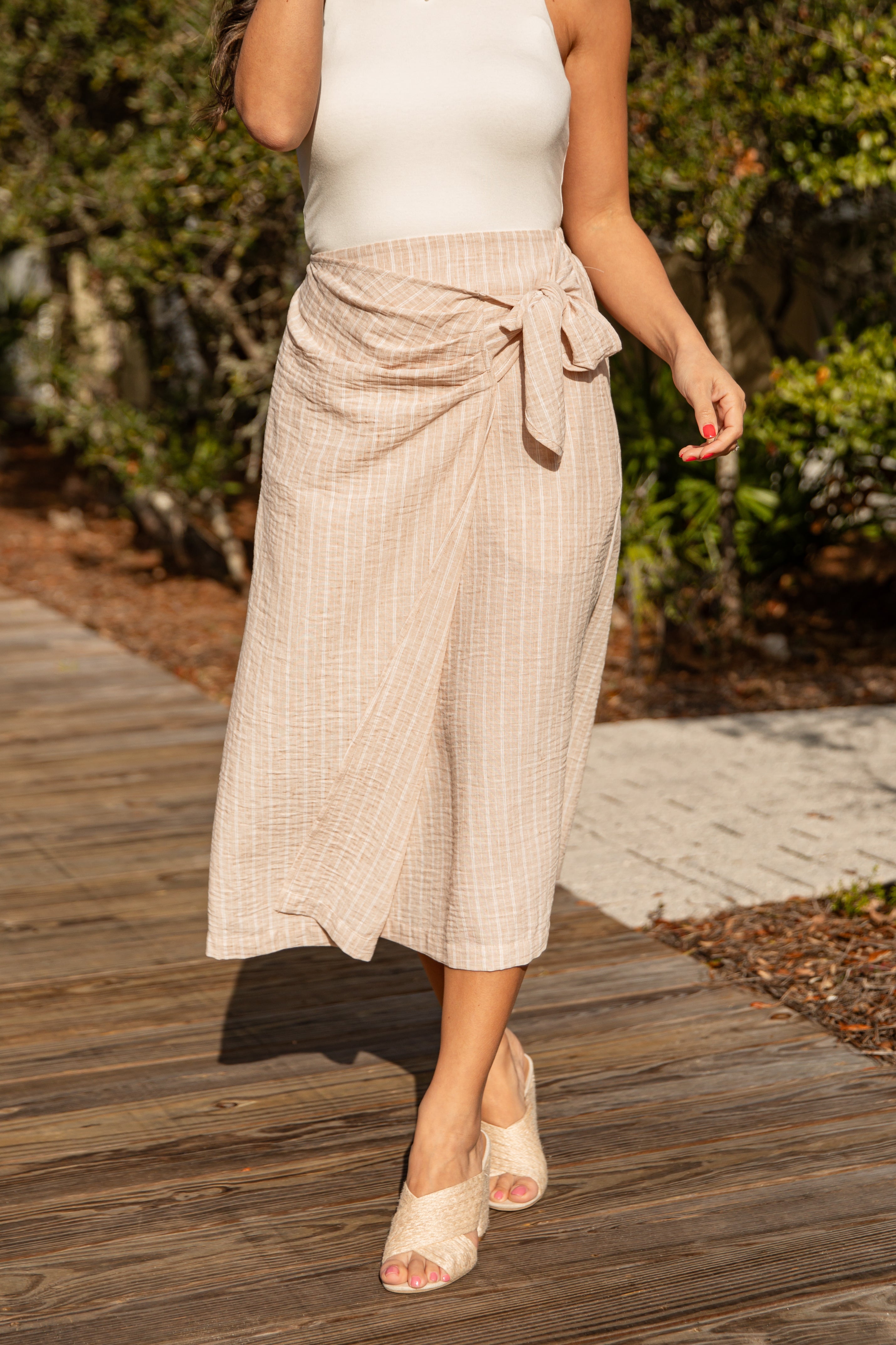 Person wearing a beige wrap skirt and white top on a wooden path with greenery.