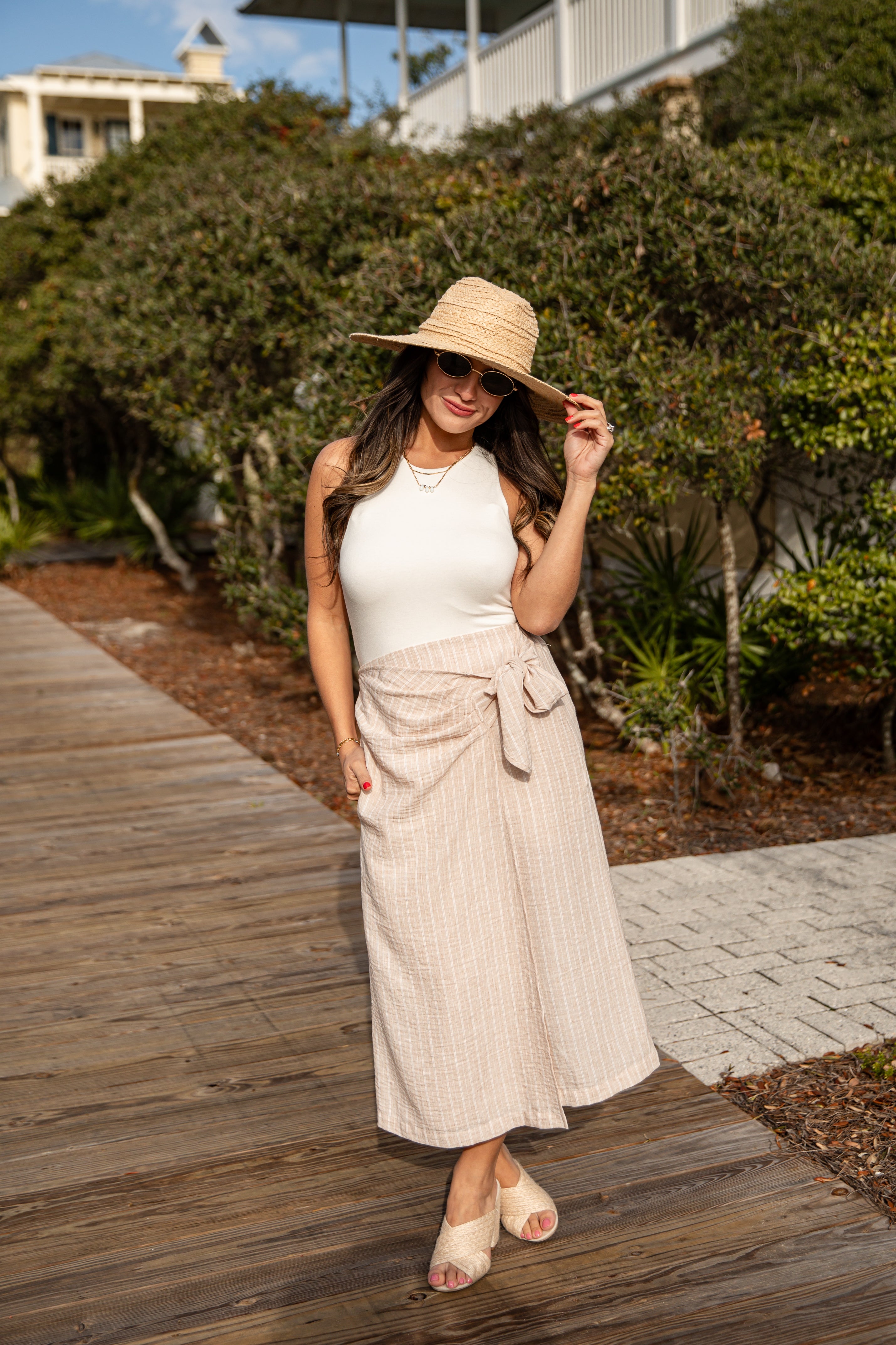 Woman in a white top and beige skirt with a straw hat standing on a wooden boardwalk.