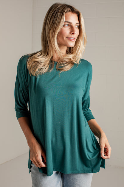 A woman wearing a teal green three-quarter sleeve top and light wash jeans, standing against a light-colored brick wall.