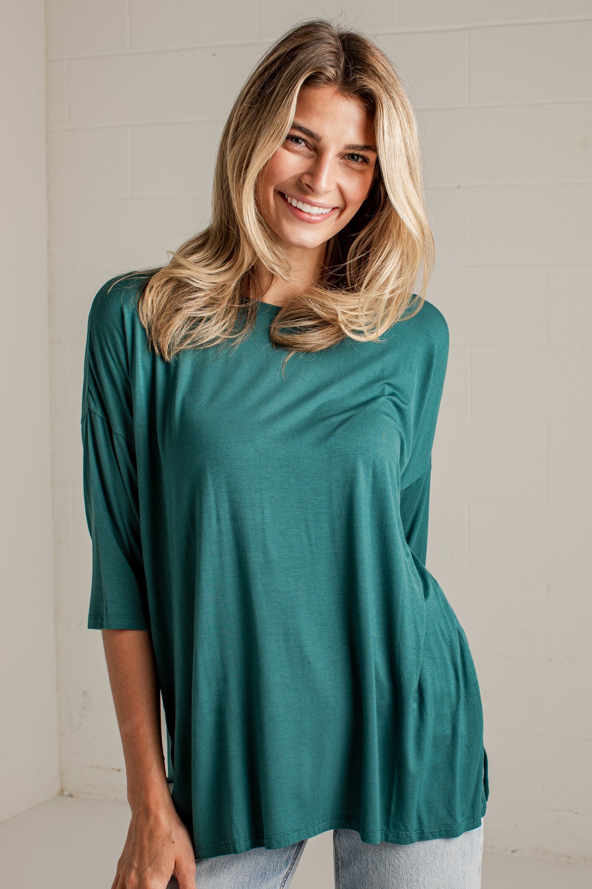 A smiling blonde woman wearing a teal green three-quarter sleeve top and light wash jeans, standing against a white painted brick wall.