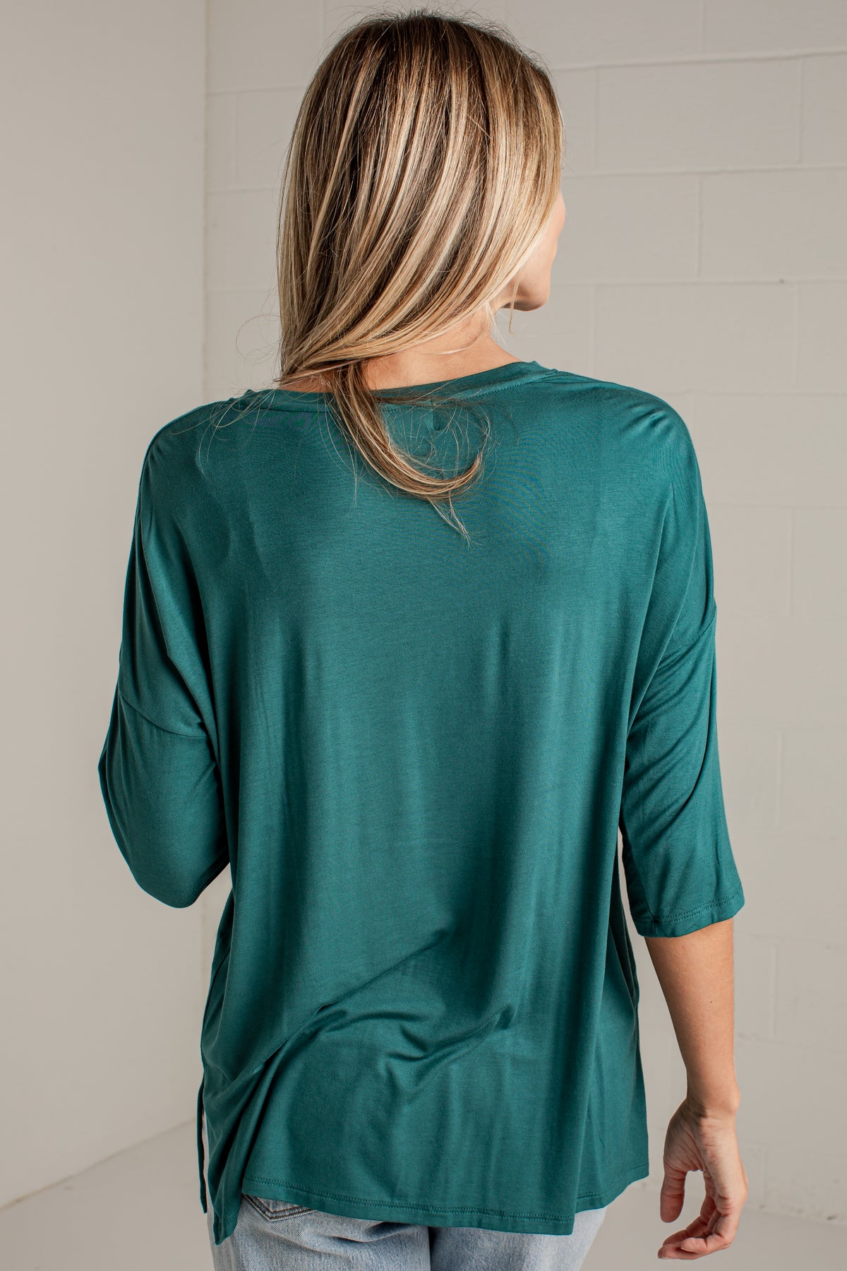 A woman viewed from behind wearing a teal green three-quarter sleeve top and light wash jeans, standing against a light gray brick wall.