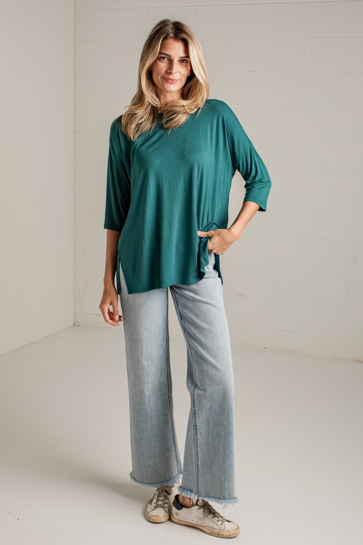 A woman modeling a teal green three-quarter sleeve top with light-wash wide-leg jeans and beige sneakers, standing in a bright, neutral studio setting.