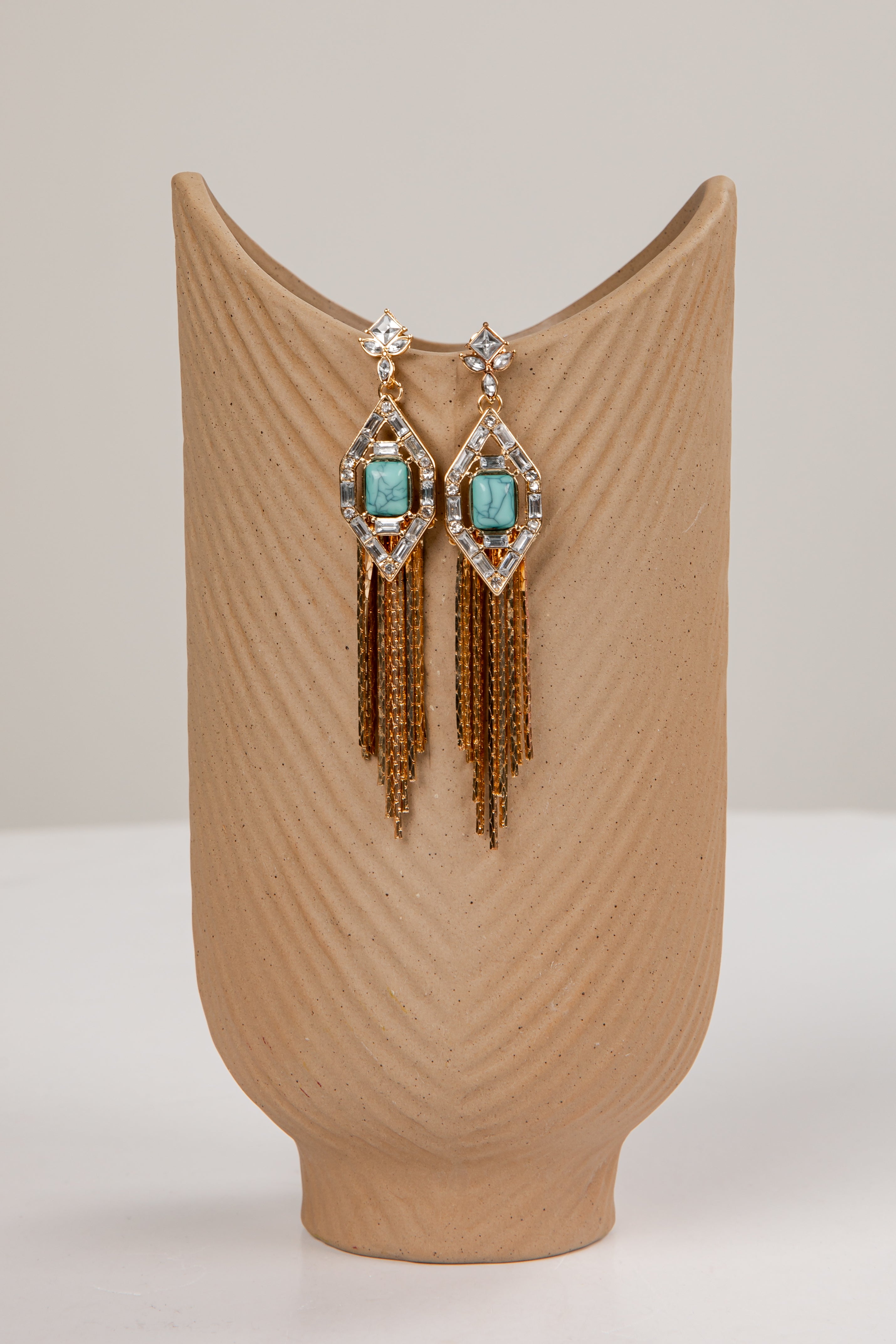 Pair of earrings with turquoise stones and gold fringes on a beige stand.