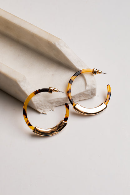 Tortoiseshell hoop earrings with gold-toned metal accents, displayed on a light gray surface next to a white stone prop.