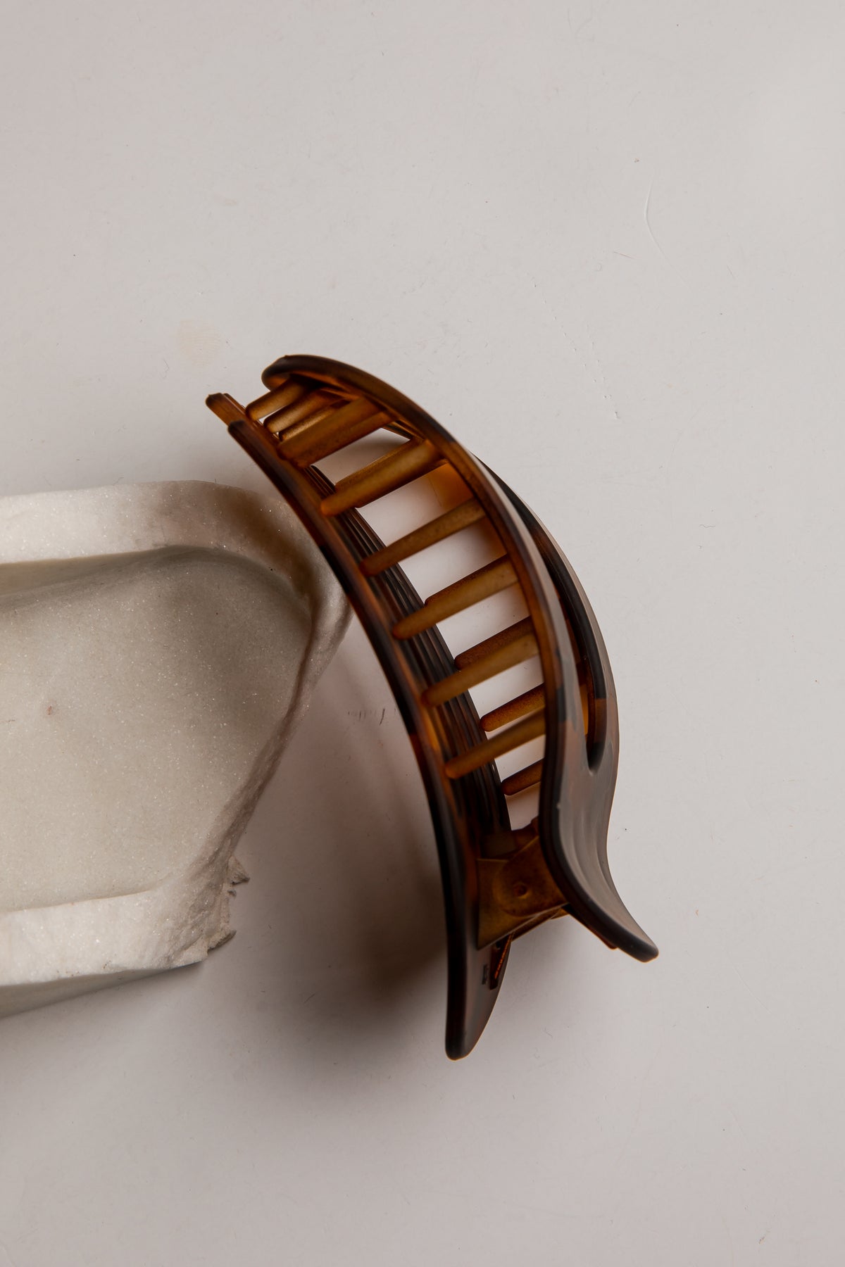 A single brown translucent hair claw clip photographed on a plain light background, with part of a white textured prop visible at the lower left.