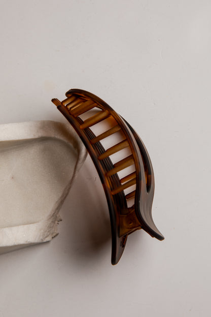 A single brown translucent hair claw clip photographed on a plain light background, with part of a white textured prop visible at the lower left.