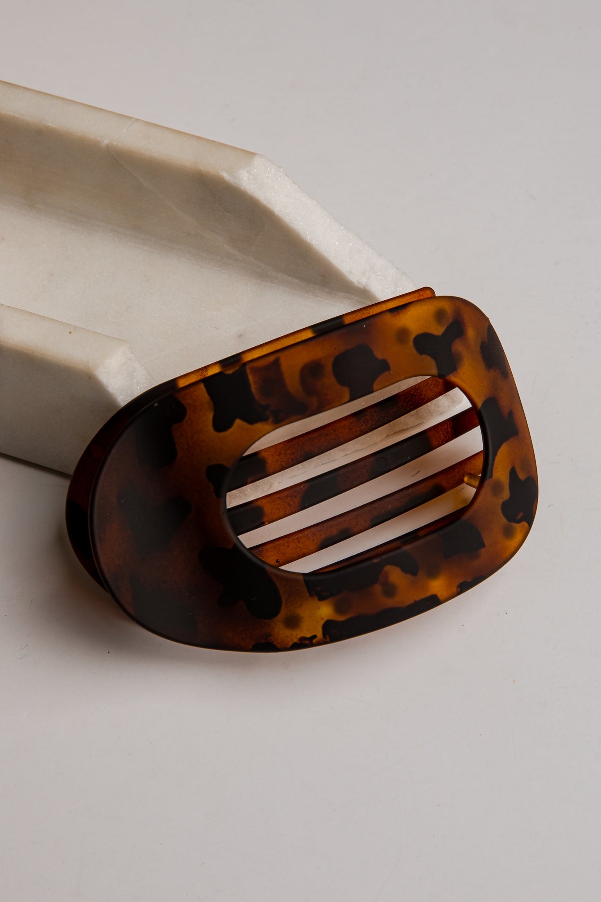 A tortoiseshell hair claw clip with a glossy amber and black pattern, resting on a white marble prop against a light gray background.