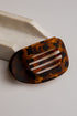A tortoiseshell hair claw clip with a glossy amber and black pattern, resting on a white marble prop against a light gray background.