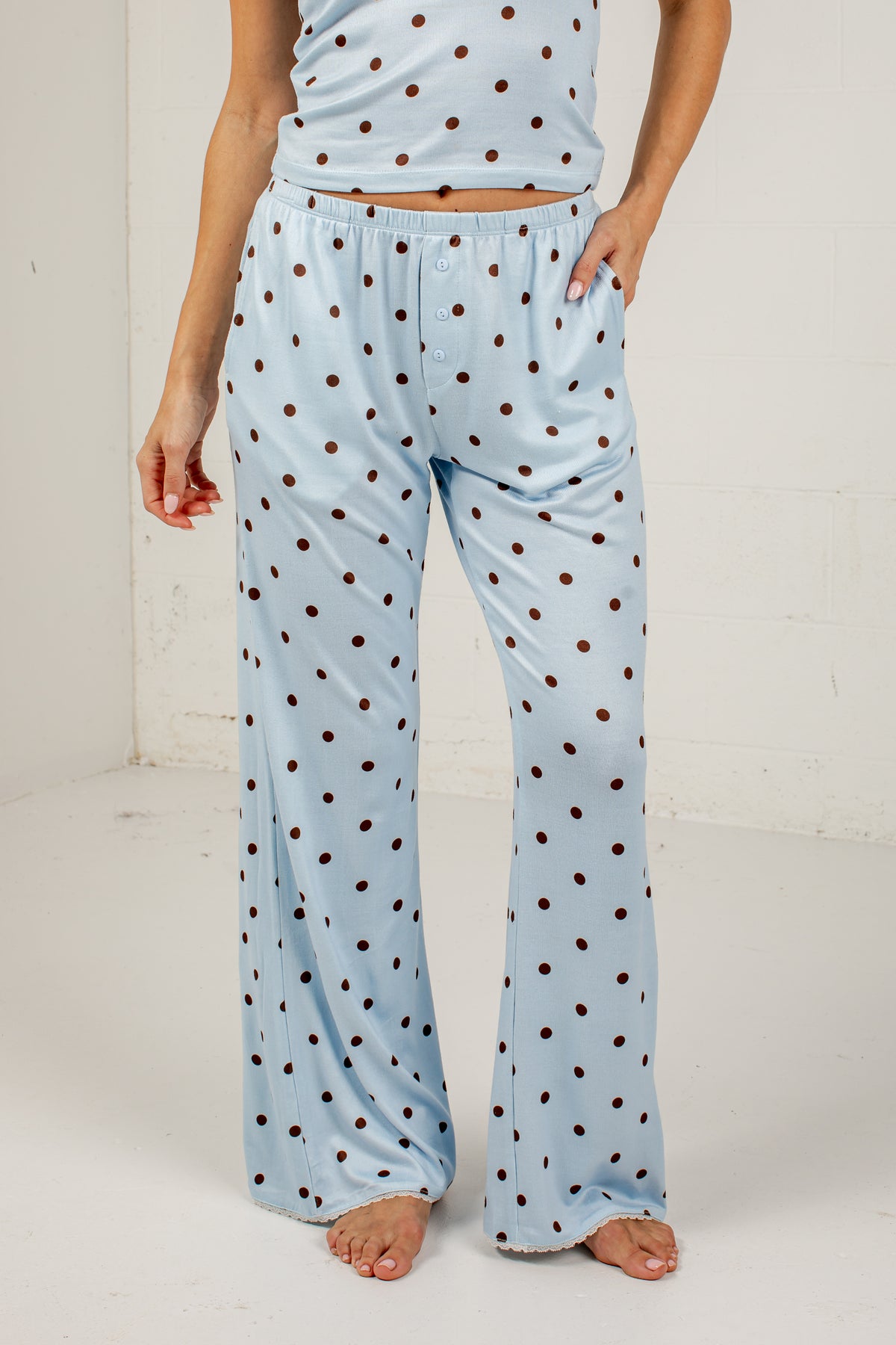 A person wearing light blue pajama pants with dark brown polka dots and a matching top, standing barefoot against a white studio wall.