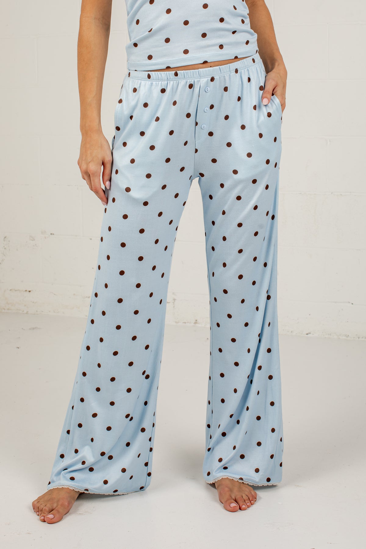 A person wearing light blue wide-leg pajama pants with a brown polka dot pattern, shown from the waist down against a white studio wall.