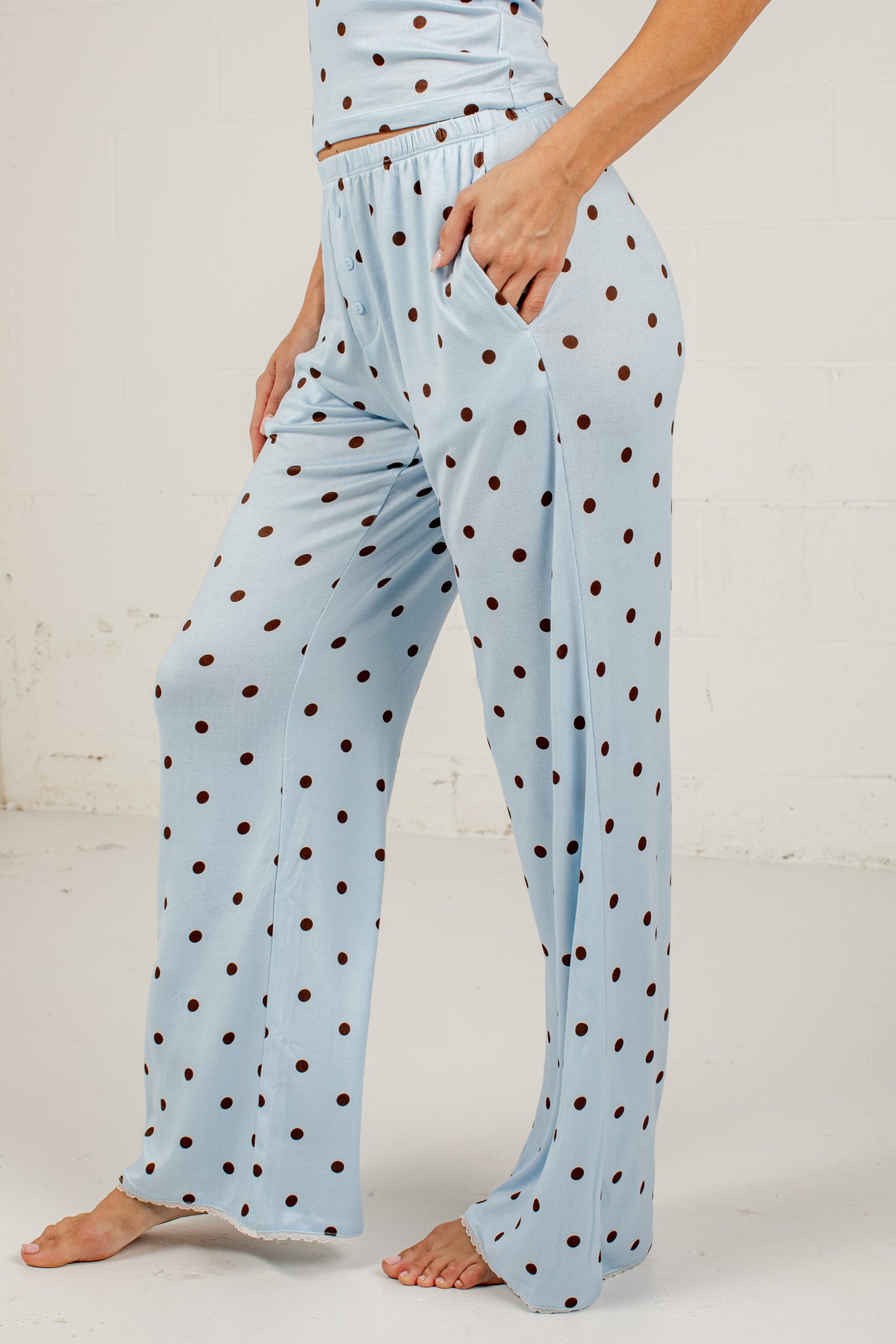 A person wearing light blue wide-leg pajama pants with dark brown polka dots, standing barefoot against a white wall.