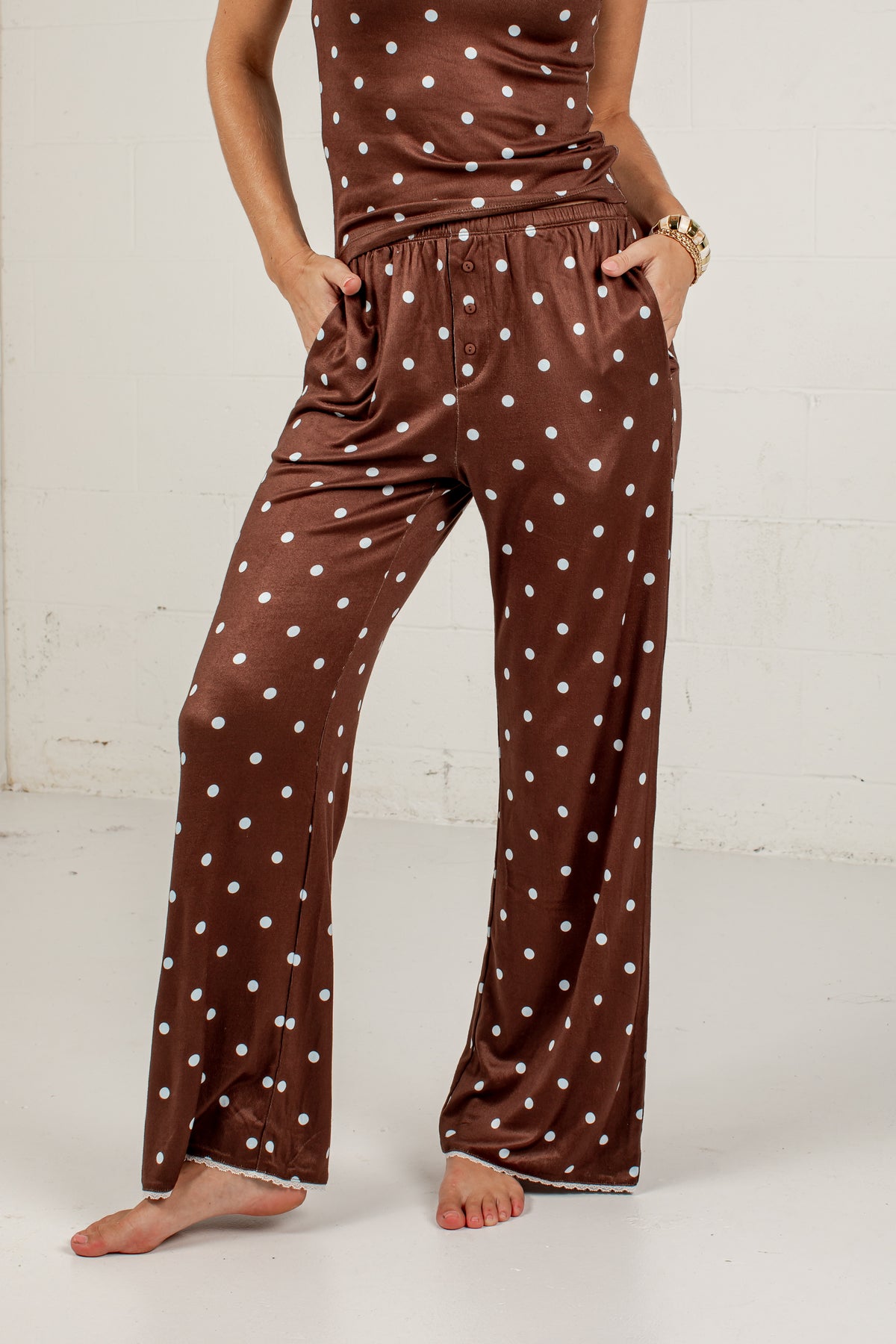 A person wearing brown satin pajama pants with white polka dots and a matching top, standing barefoot against a white wall.