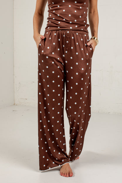 A person wearing brown wide-leg pants with a white polka dot pattern, standing barefoot against a white studio wall.