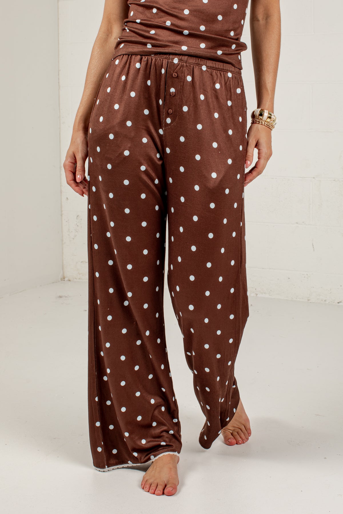 A person wearing brown wide-leg pajama pants with a white polka dot pattern, shown from the waist down in a studio setting.