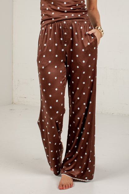 Brown wide-leg pants with a white polka dot pattern, shown on a model standing barefoot against a white wall.