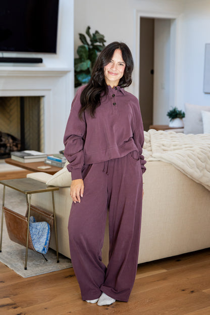 A woman wearing a matching mauve-purple lounge set consisting of a long-sleeve button-front top and wide-leg pants, standing in a cozy living room with wood floors, a bed, and a fireplace.