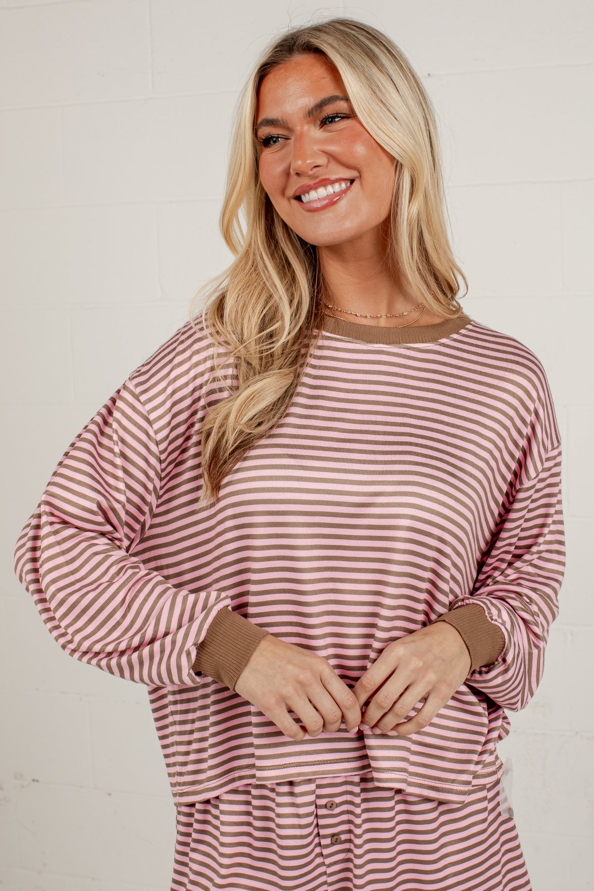 A smiling blonde woman wearing a long-sleeve dusty rose and white striped top with solid brown ribbed cuffs and collar, standing against a white brick wall.