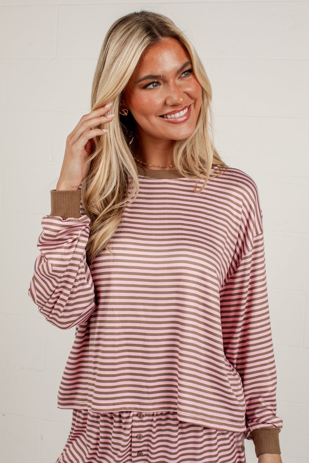 A smiling blonde woman wearing a long-sleeve dusty rose and white striped top with brown ribbed cuffs, standing against a light off-white brick wall.