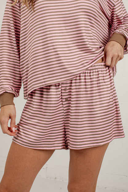 A close-up of a person wearing a matching two-piece loungewear set with horizontal pink and white stripes, featuring a long-sleeve top and shorts with brown ribbed cuffs and waistband.