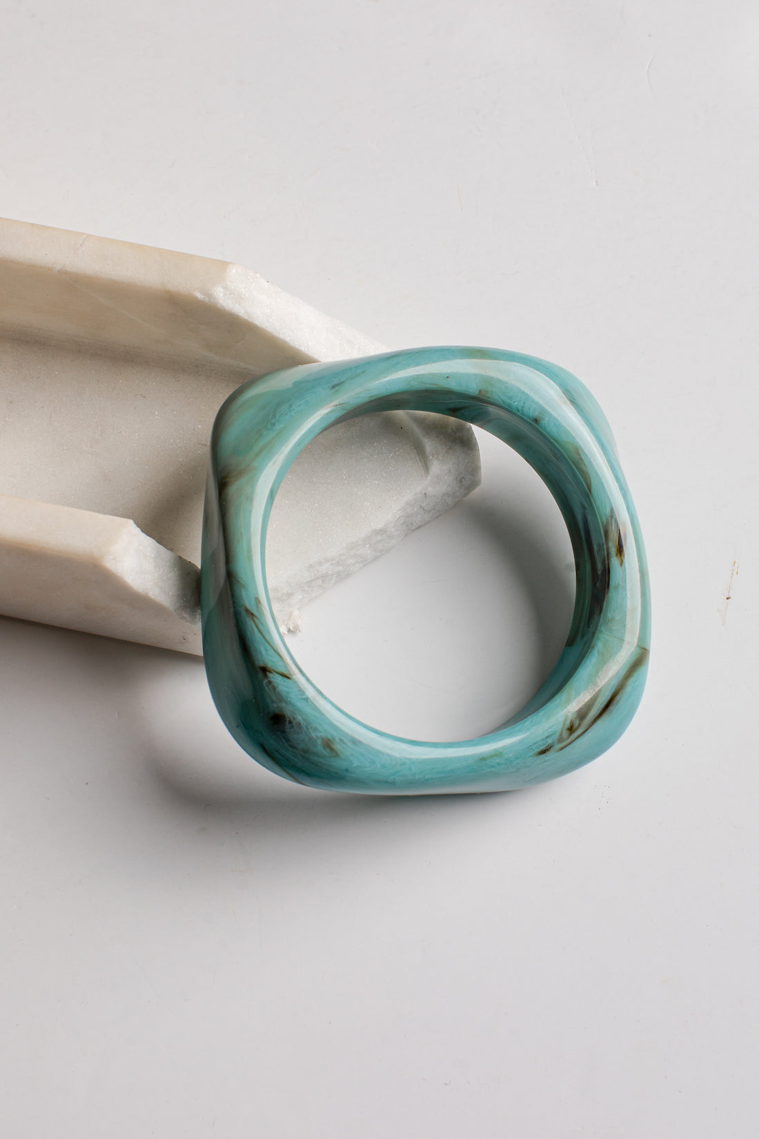 A chunky square ring made of turquoise stone with natural brown and dark veining, displayed on a light gray surface next to a white geometric prop.