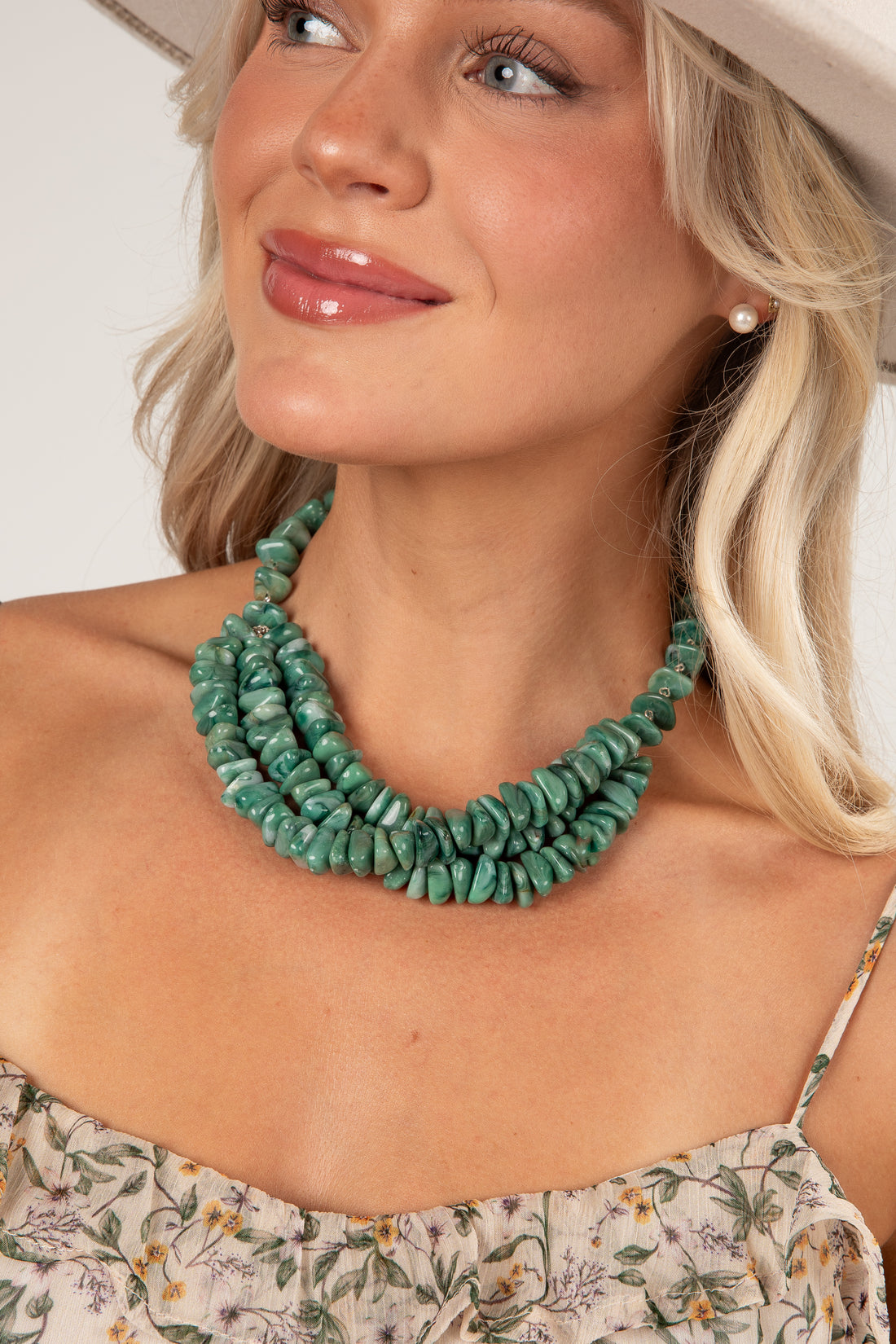 A close-up of a woman wearing a chunky, multi-strand turquoise green stone necklace, styled with a floral top and a light-colored hat.