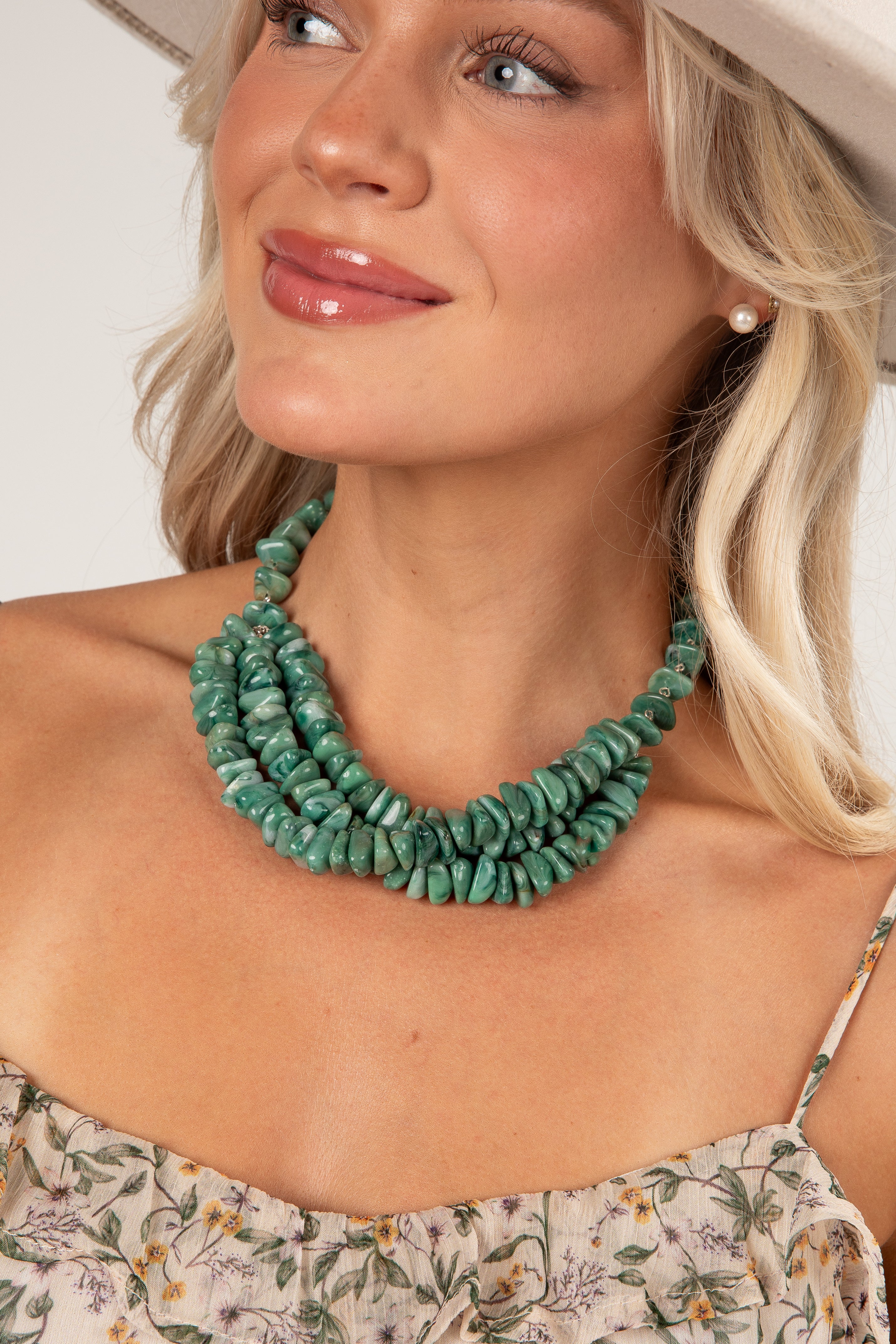 A close-up of a woman wearing a chunky, multi-strand turquoise green stone necklace, styled with a floral top and a light-colored hat.