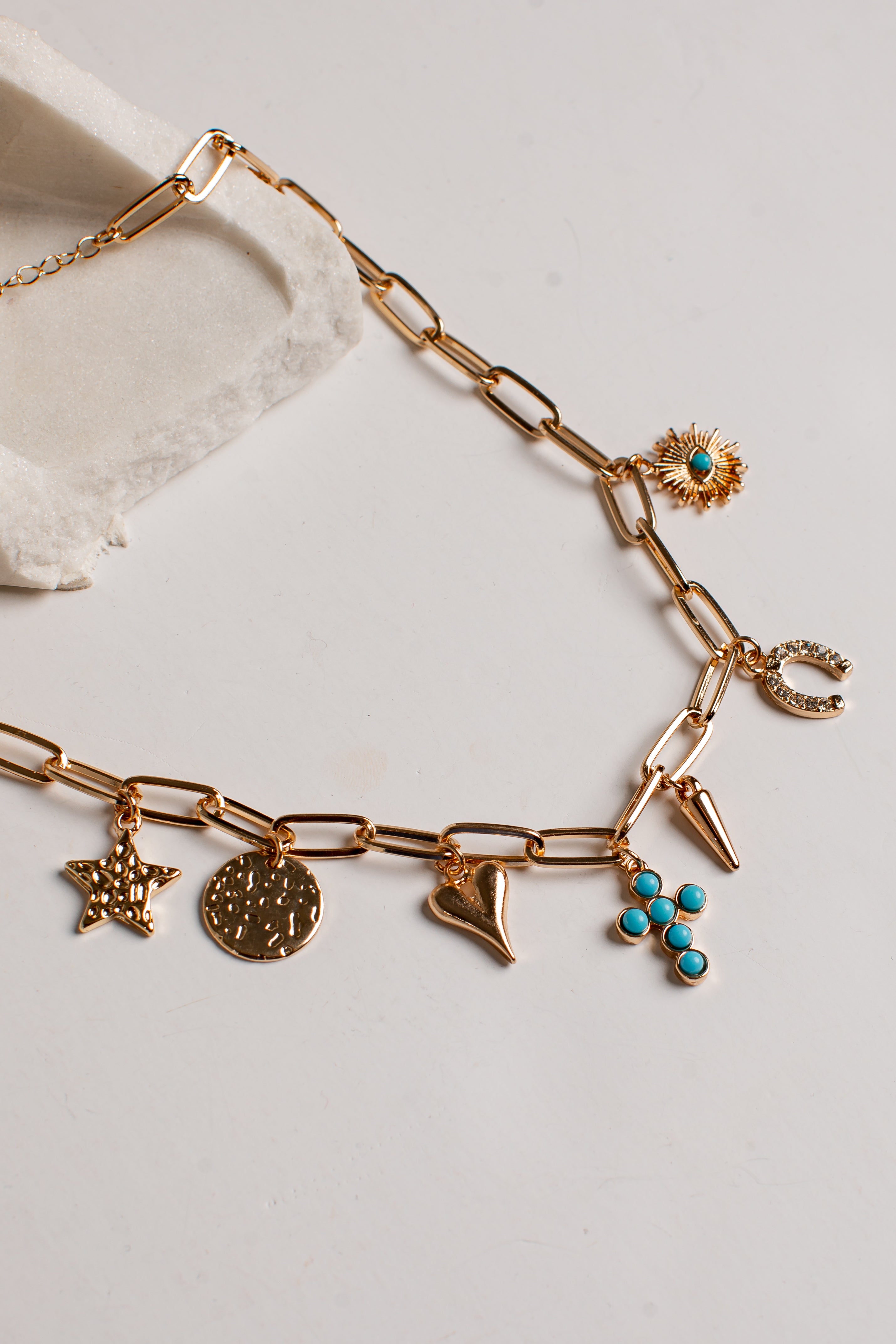 A gold-tone chain necklace with multiple charms, including a starfish, a round medallion, a crescent moon, a flower with turquoise accents, and a small sunburst charm, displayed on a light neutral surface.
