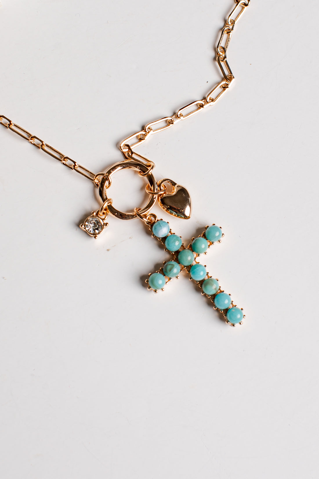 A gold-tone cross pendant necklace adorned with turquoise-colored stones, featuring a small heart charm and a clear crystal charm on a white background.