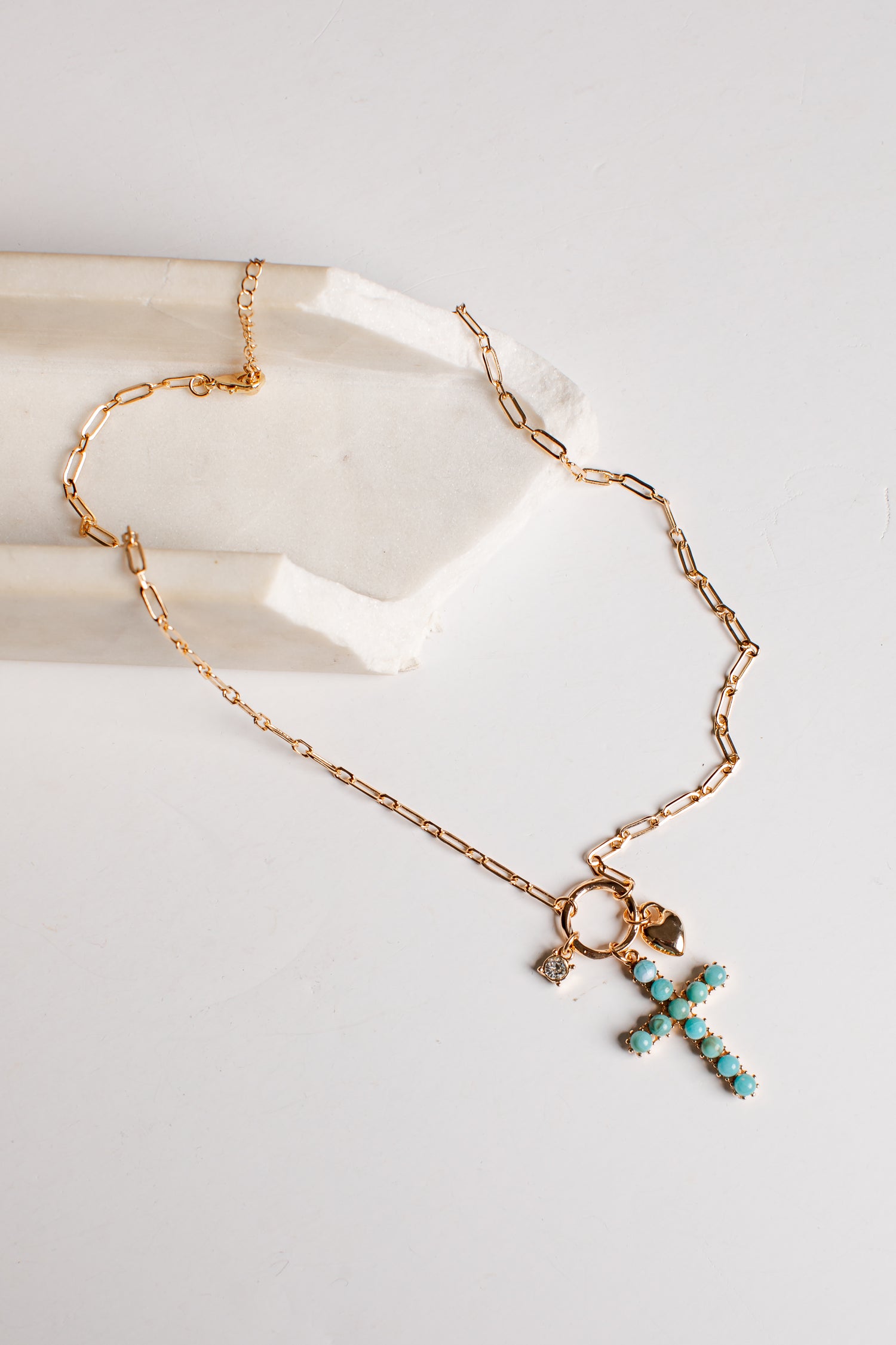 A gold chain necklace with a cross-shaped pendant studded with turquoise blue beads, displayed on a white surface next to a white stone block.