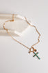 A gold chain necklace with a cross-shaped pendant studded with turquoise blue beads, displayed on a white surface next to a white stone block.
