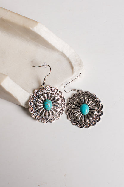 A pair of silver-toned dangle earrings with intricate floral filigree detailing and a central turquoise stone, displayed on a light cream textured surface.