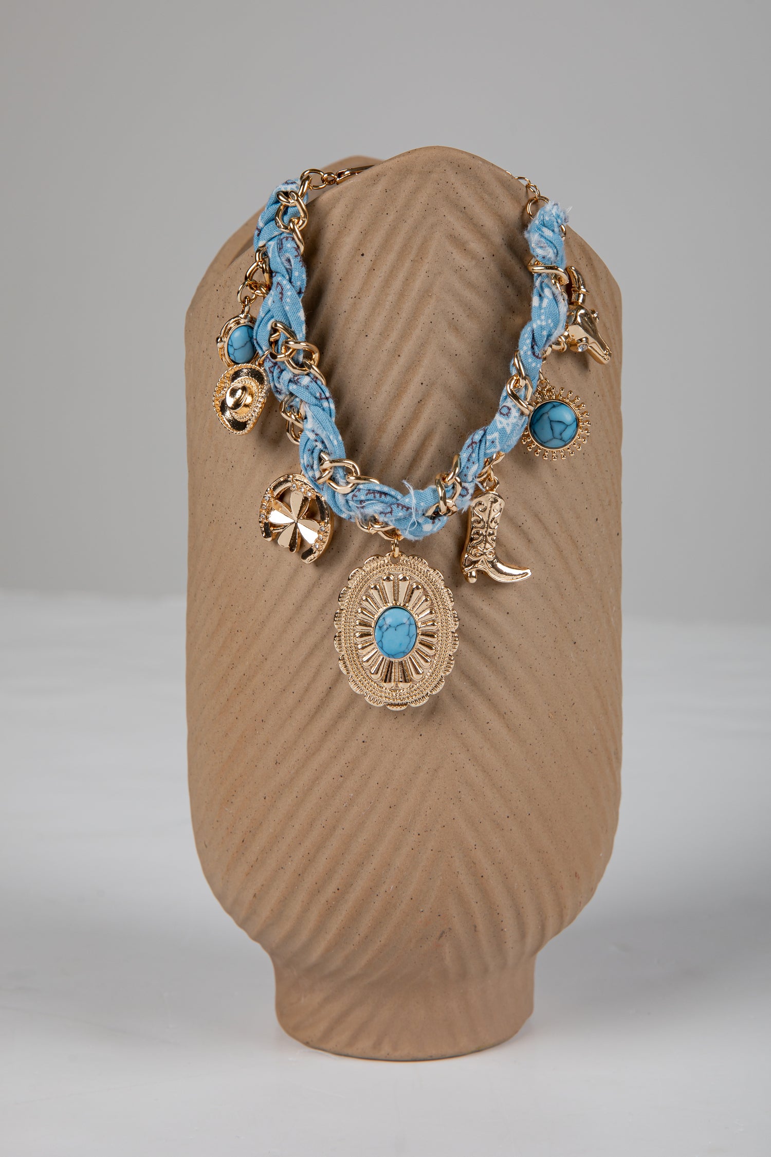 Necklace with blue braided cord and gold charms on a brown stand against a gray background