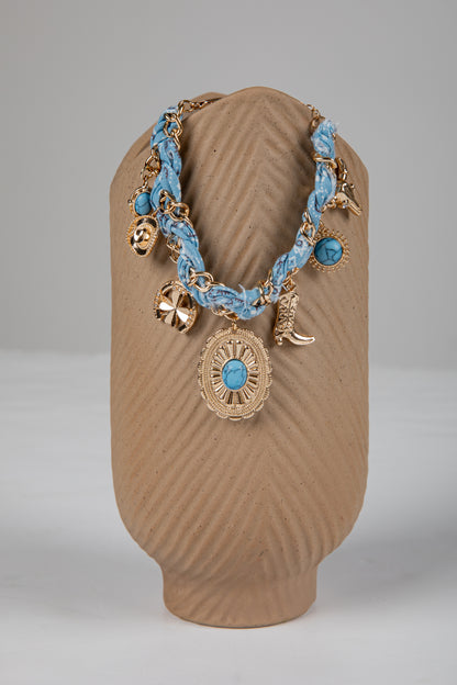 Necklace with blue braided cord and gold charms on a brown stand against a gray background