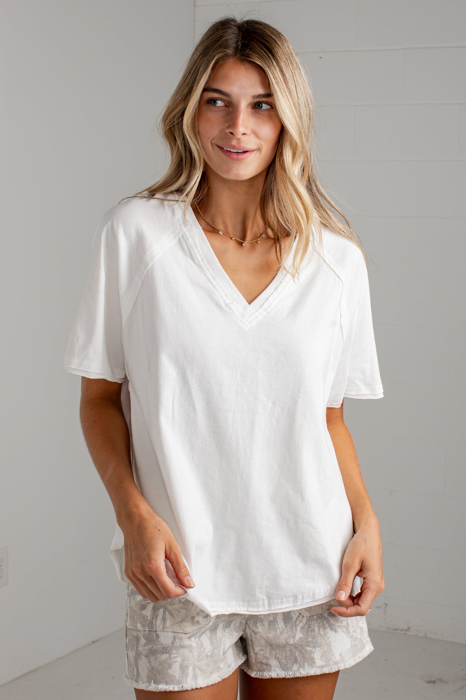 A woman wearing a white short-sleeve V-neck t-shirt and light gray patterned shorts, standing against a light gray wall.