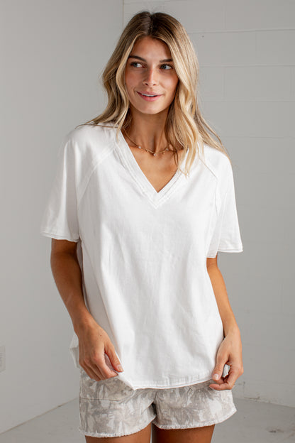 A woman wearing a white short-sleeve V-neck t-shirt and light gray patterned shorts, standing against a light gray wall.