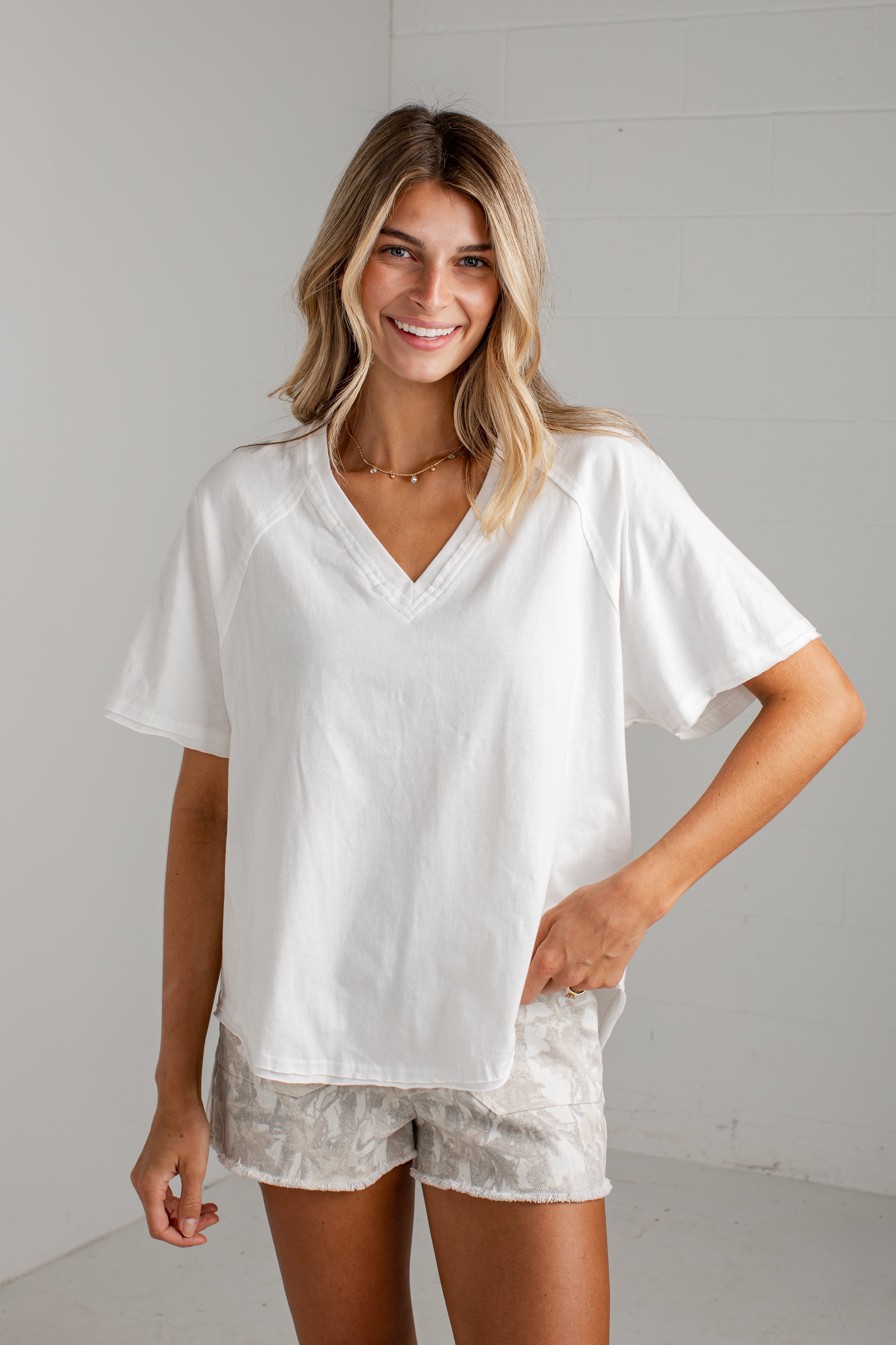 A woman modeling a white short-sleeve V-neck top with a relaxed fit, paired with light patterned shorts, standing against a light gray wall.