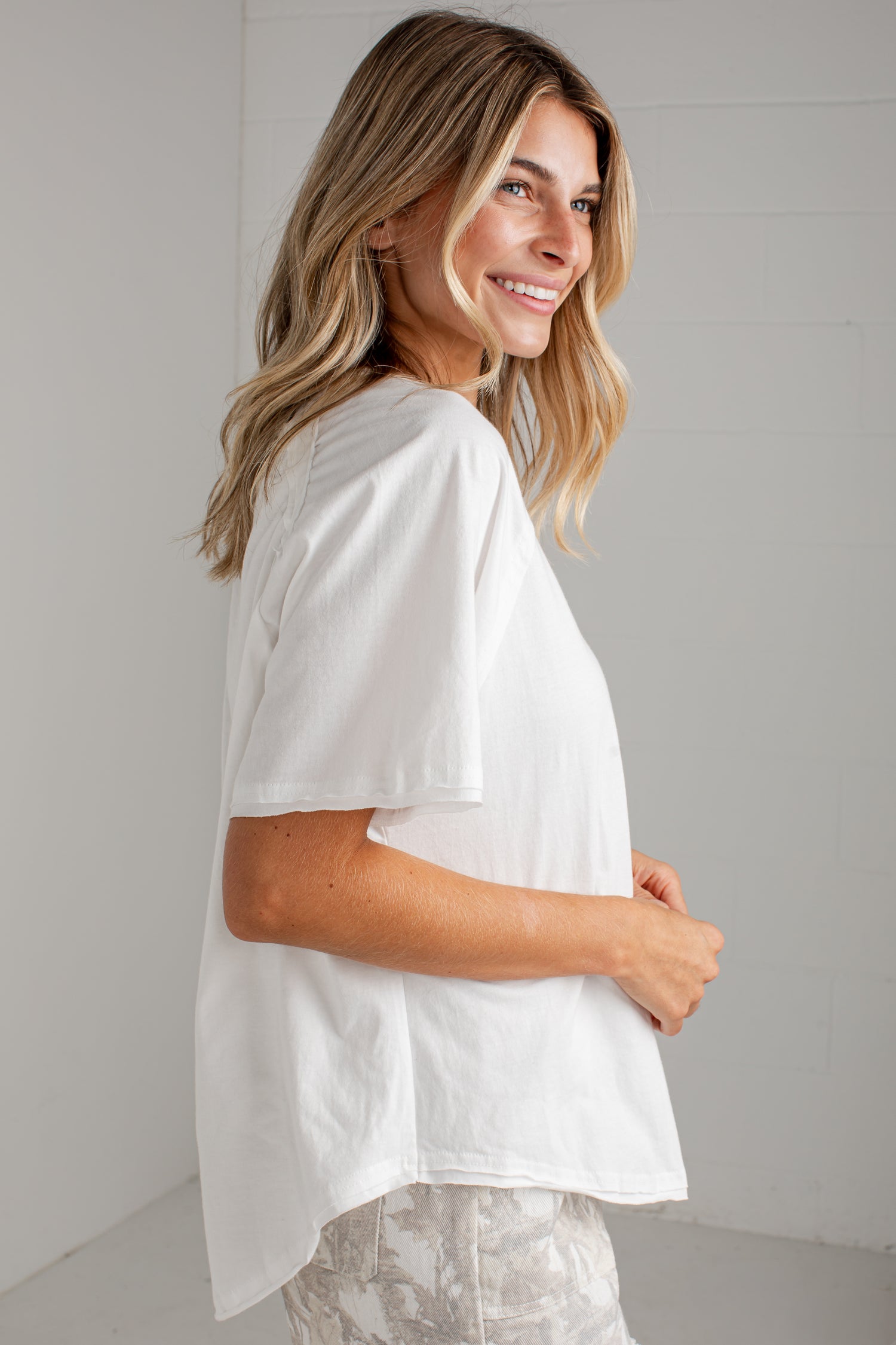 A woman wearing a loose white short-sleeve top and light patterned pants, photographed in a bright indoor setting with a white wall background.