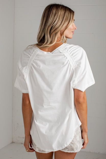 A woman shown from the back wearing a loose white short-sleeve top with decorative shoulder straps and a lace-trimmed hem, standing against a white paneled wall.