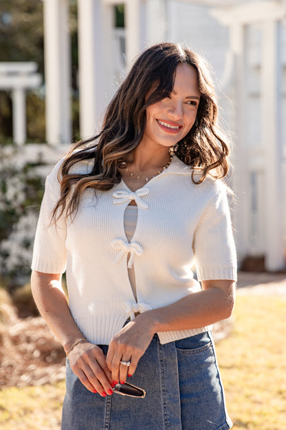 Woman wearing a white short-sleeve top with a bow detail, standing outdoors.